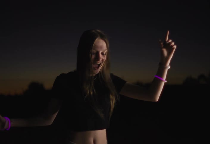 A young woman dancing and enjoying herself outdoors at dusk or nightshe is wearing a dark crop top and has long hair that moves as she dances and she is accessorized with several glowing bracelets on her wriststhe background is very dark with only