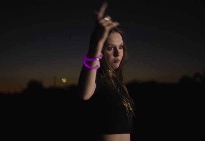 A woman dances and sings in slow motion at dusk lit by a flashing light source and wearing glowing wristbands