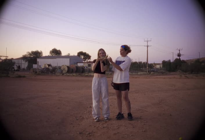 Two women look at a phone on a dirt lot at sunset
