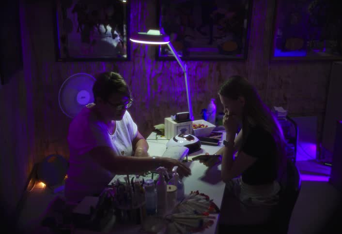 A client chooses nail designs with a manicurist under purple light in a dim room