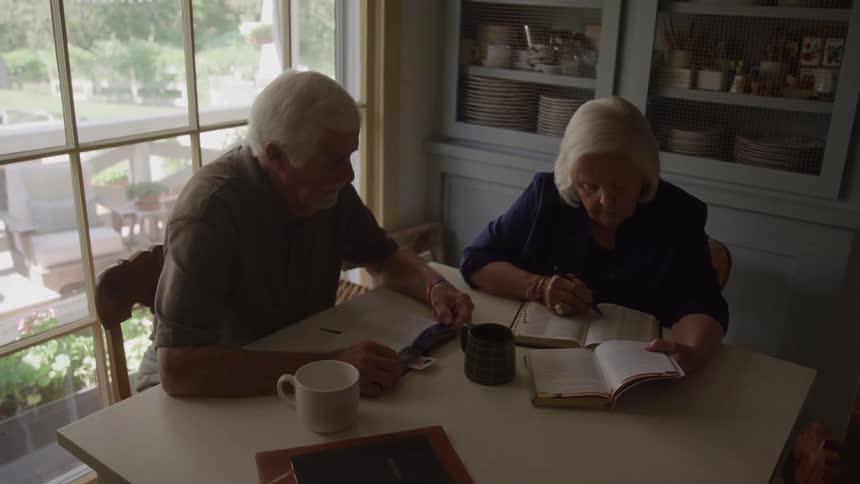 An older man and woman are sitting together at a white table reading books and writing in notebooks with coffee mugs nearby in a brightly lit room with a large window
