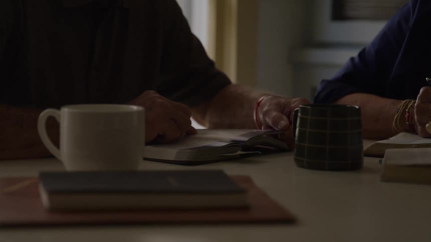 Hands two elderly people reading a book and making annotations over a table indoors during day time