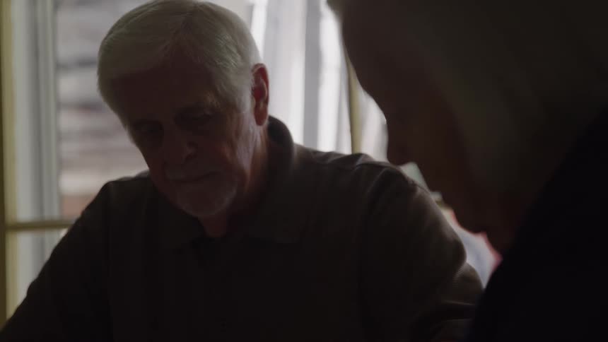 The video is a closeup shot of two older adults one in the foreground facing away from the camera and the other a man with white hair facing down and slightly toward the camerathe man appears to be intently focused on something below him possibly