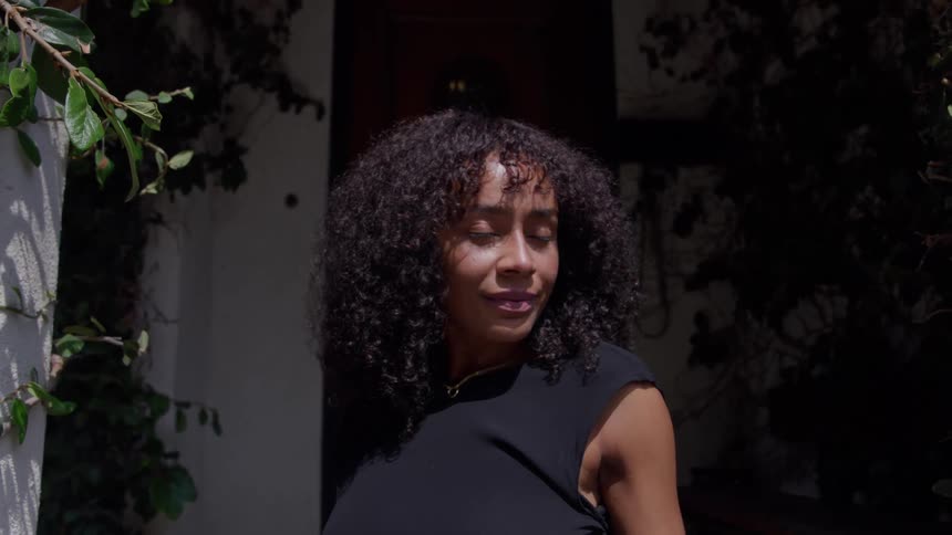A closeup shot features a young woman with dark voluminous curly hair she is standing outdoors in a shaded area wearing a black top and looks directly at the camera with a calm expression