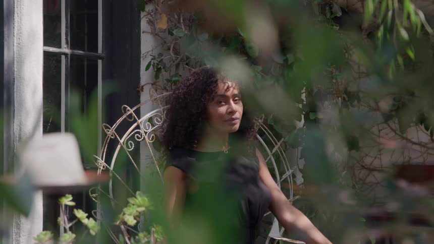 The video shows a woman with curly hair sitting on an ornate white metal chair outdoorsthe shot is framed through outoffocus green foliage with the woman looking directly towards the camera