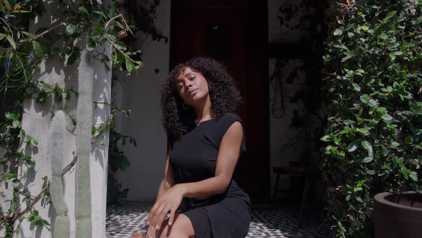 A stylish young woman with dark curly hair wearing a sleeveless black dress sits on a patterned tiled porch basking in bright sunlight between two walls covered in lush green vines