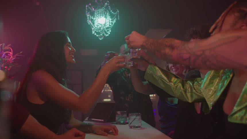 A happy group of friends raise their shot glasses for a toast over a white bar counter beneath a glowing chandelier in a dimly lit neoncolored nightclub