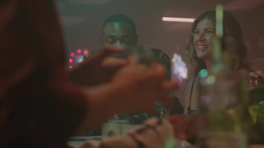 A happy man in a dark patterned shirt and a smiling woman in a blue dress sit together at a brightly lit bar counter