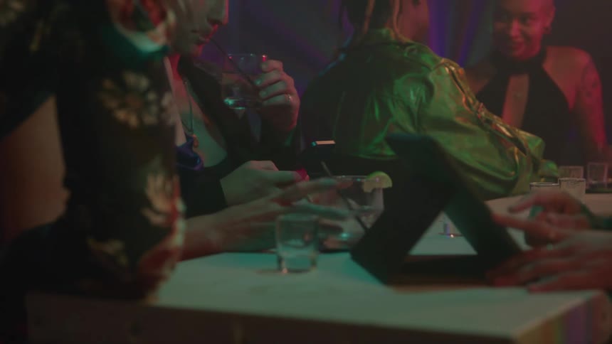 The video shows a group of people sitting at a table in a dimly lit club or bar with the scene illuminated by neon or colored lightspeople are dressed in party attire and appear to be socializing with drinks and glasses visible on the tab