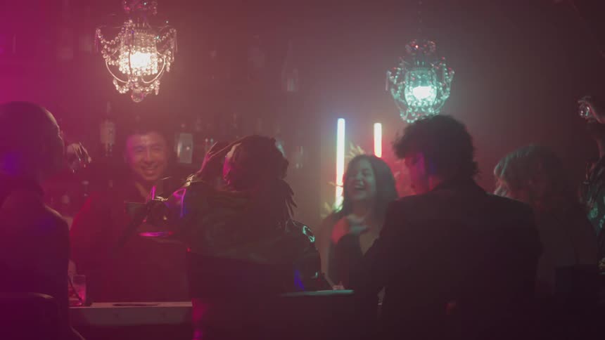 The video shows a lively dimly lit scene inside a nightclub or bar with several people gathered around a bar or hightop tablethe atmosphere is characterized by colored lighting pink blue and green and two elaborate chandeliers hanging overhead