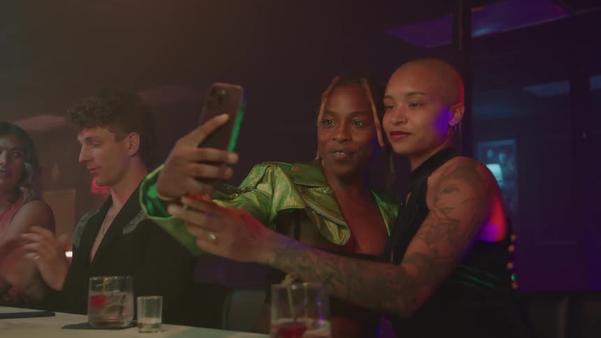 The video shows two people in a dimly lit clublike setting taking a selfie together using a smartphonethey are seated at a table or bar smiling and posing for the picture with other patrons visible in the background