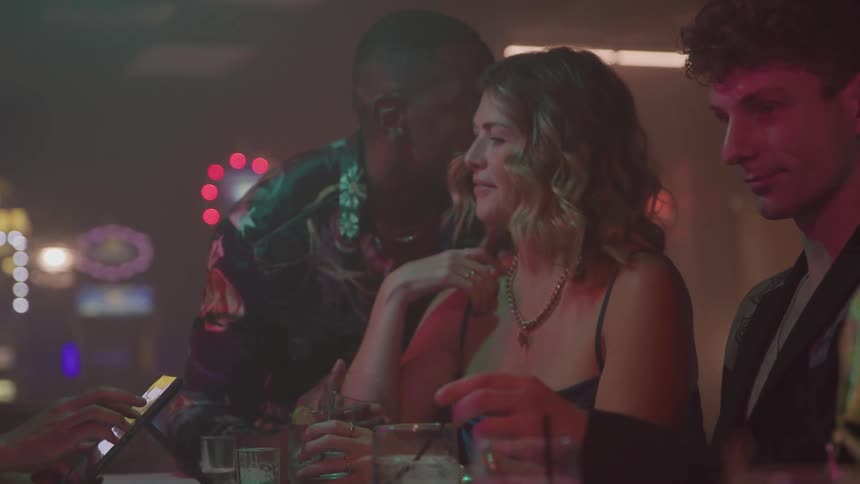 A group of friends is gathered at a bar or club setting illuminated by the venues colorful mood lighting they are socializing smiling and enjoying drinks in a lively nightlife atmosphere