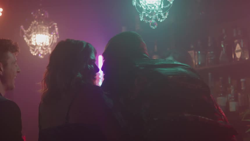 A man and a woman stand close together kissing in a dimly lit nightclub with two chandeliers while another man stands near a wells tocked bar in the background