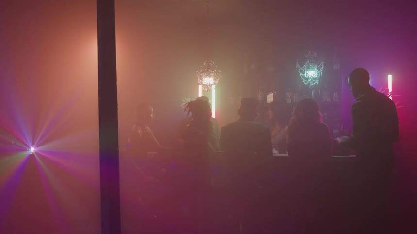 The scene shows several people gathered in a dimly lit atmospheric space like a bar or club flashes of light including a vibrant sweeping laser effect on the left add to the dark hazy ambiance