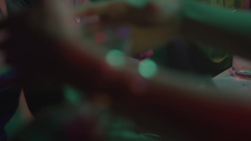 A group of friends sit around a dining table at night engaged in lively conversation while one of them quietly texts on her phone