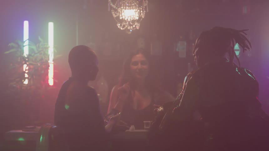 Two women sit across a bar counter from a bartender wearing a low cut top talking in a smoky dimly lit club illuminated by colorful vertical neon lights and a hanging crystal chandelier