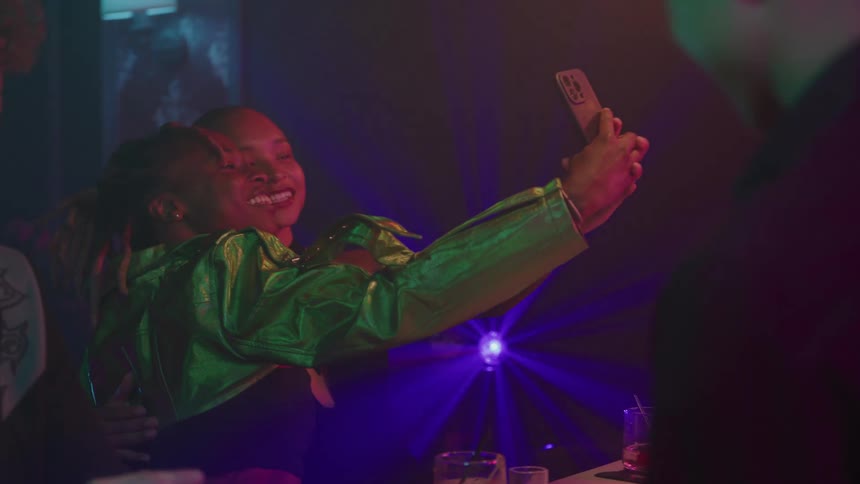 Two young smiling women take a selfie with a phone one wearing a green metallic jacket and the other with dreadlocks kissing her cheek in a dark club illuminated by disco lights