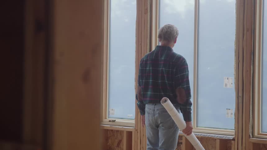 A middleaged construction engineer inside a building under construction inspecting the windows while holding a rolledup building plan on his arm at midday