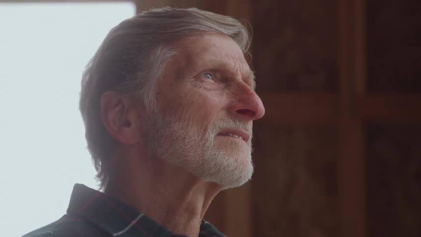 A tight profile shot focuses on the face of an elderly man with gray hair and a short white beardhe is looking up and away from the camera bathed in soft light coming from an unseen window or opening