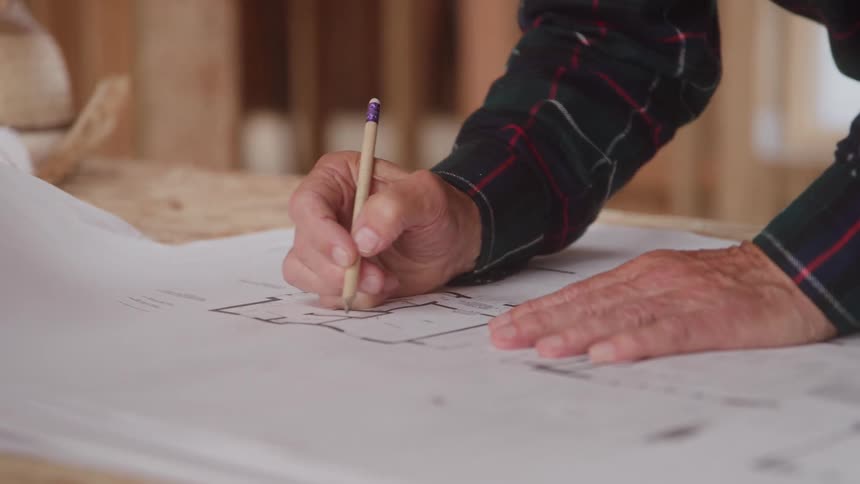 An architect drawing a building plan on white paper at a desk inside a building under construction at midday
