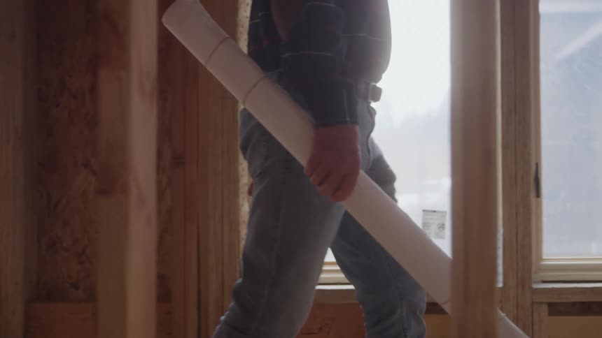 A construction worker dressed in a dark shirt and jeans walks across the frame inside a room under constructionthe worker is carrying a large roll of blueprints or building plans and walks past a new window letting in bright daylight