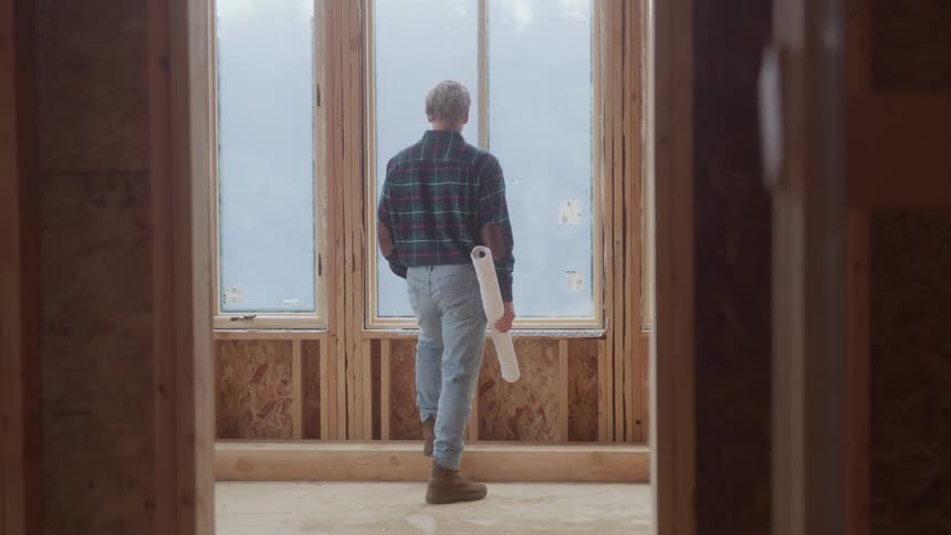 A worker wearing a plaid shirt and jeans stands facing a tall window in a house under construction holding a rolled up blueprint while viewing the empty space and wooden wall studs