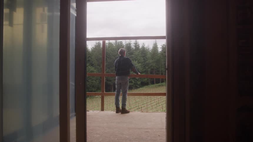 A profile view shows a mature bearded worker wearing a plaid shirt looking thoughtfully from the balcony of a house under construction