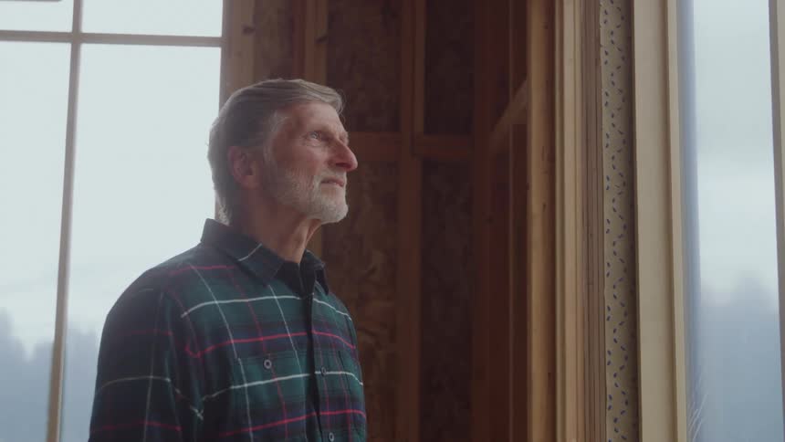 A profile view shows a mature bearded worker wearing a plaid shirt looking thoughtfully out of a large window inside a room of a house under construction