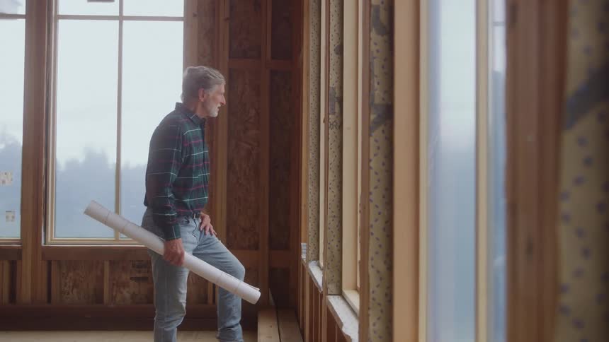 A middle aged man walks towards a window inside a building under construction in daytime