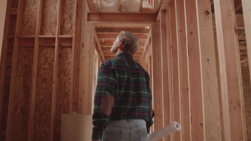 A middleaged man holding an architectural drawing walks through a building under construction inspecting the wooden structures as he moves at midday