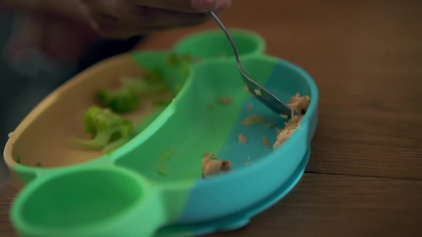 A person sitting at a dining table feeding a baby from a plate with a spoon