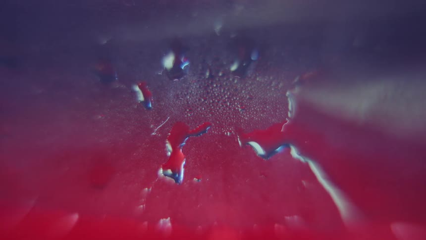 Close up drops slide down a steamed glass surface creating a unique texture over a background of dark purple and bright red color gradients