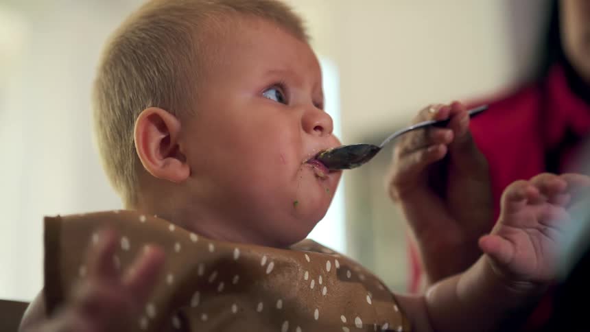 A baby wearing a dotted bib is fed with a spoon while gazing sideways and reaching a small hand towards a female adult