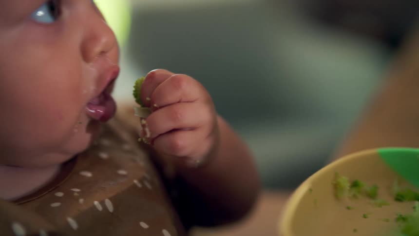 A baby eating with her hands while sitting in the dining area at midday