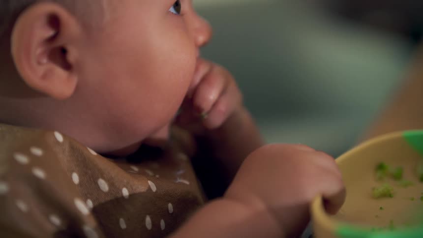 The baby holds the bowl while eating broccoli