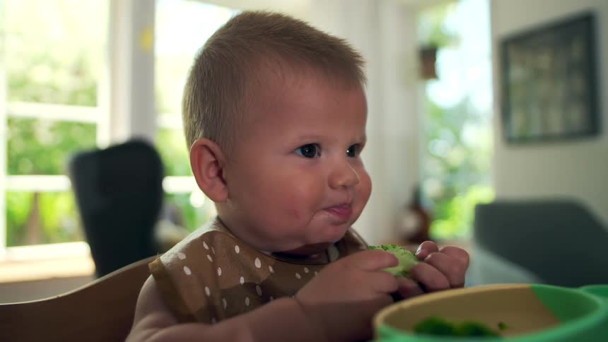 A toddler eats sliced cucumber while sitting in a high chair in a bright kitchen