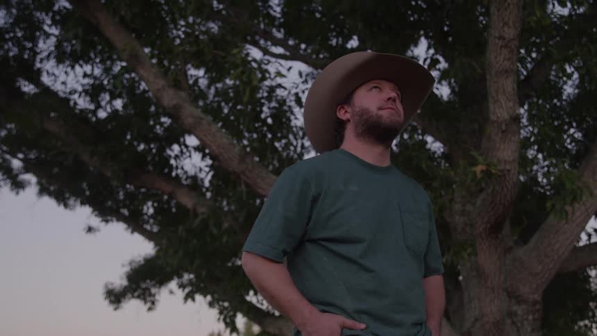 A man wearing a cowboy hat green tshirt and jeans stands outdoors against the dark silhouette of a large leafy tree at dusk looking upwards and slightly offcamera with a contemplative expression while keeping his hands in his pockets