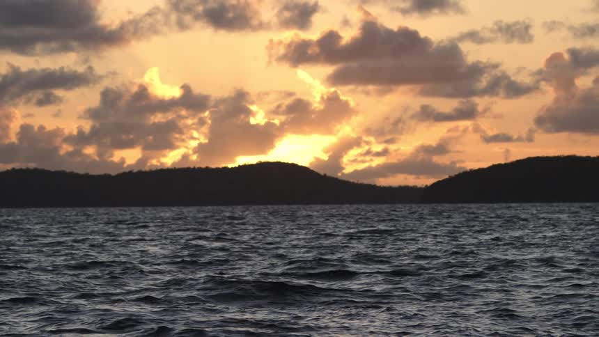 Dark choppy ocean waves roll as the sun sets behind a silhouetted island casting a brilliant golden glow across the cloudy sky