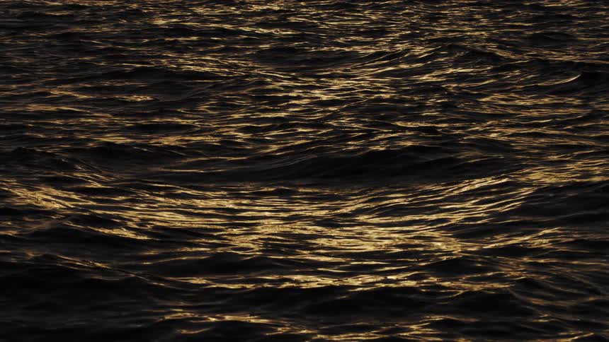 Dark choppy ocean waves roll in slow motion as the surface glitters with the golden reflections of a setting sun