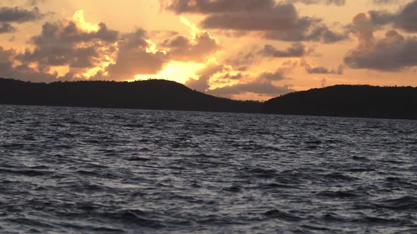 The setting sun cast a brilliant fiery glow over the jagged cloudstrewn horizon silhouetting the dark mountainous landmass the choppy deep blue water of the bay was alive with restless shimmering waves beneath the dramatic sky
