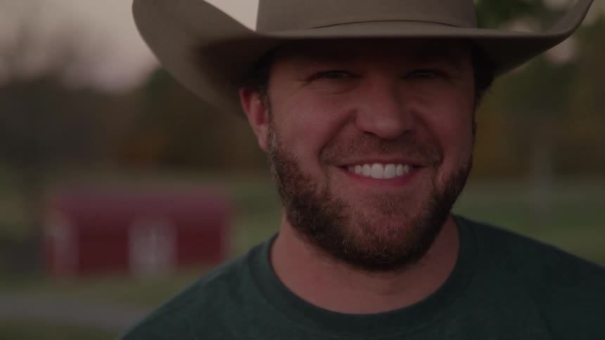 A young man with a bearded face wearing a cowboy hat on a ranch at midday