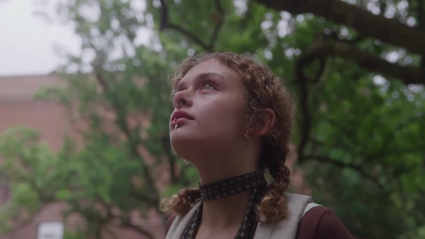 The face of a young girl with her hair tied up in braids is looking upwards and she has several piercings in her mouth and nose