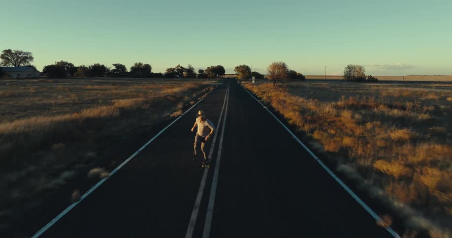 A white male skateboards down the center of a rural highway at sunset