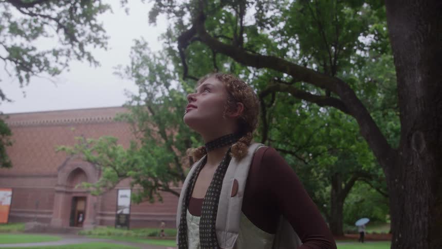 A closeup shot of a young woman standing outdoors looking up towards the sky and trees behind her is a large redbrick building which appears to be a museum or large institutional building partially visible through the trees the surrounding area is