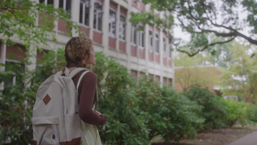 A girl wearing a white backpack is walking along a path near some vegetation and a building