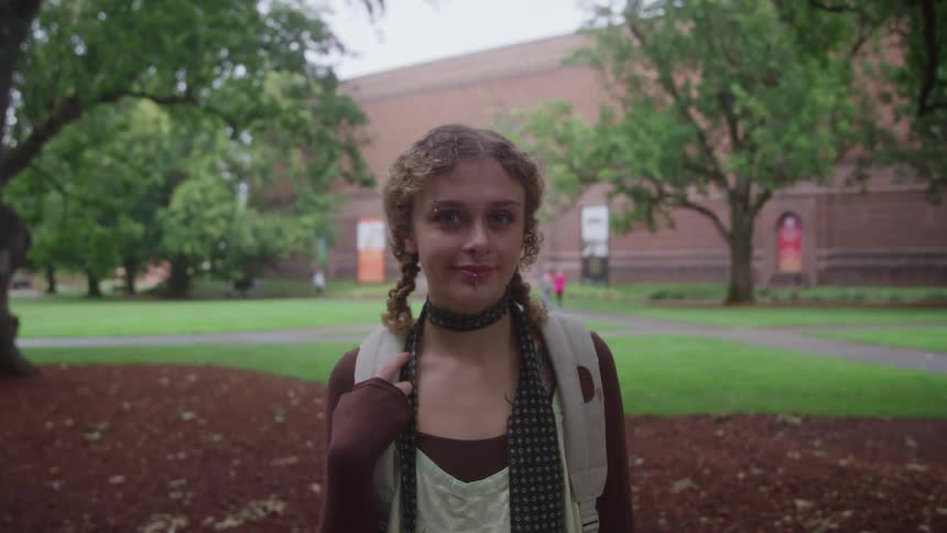 A teenage girl with curly hair styled in pigtail braids wearing a backpack and a patterned scarf stands in acollege campus looking directly at the camera with a large reddishbrown brick building visible clearly in the background
