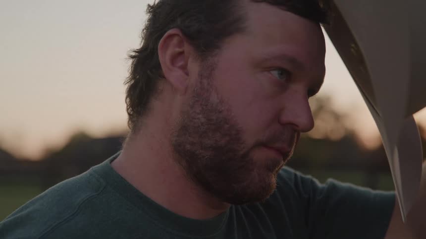 A closeup of a bearded man initially shows him with a troubled expression before he covers his face with his hand in distress he then puts on a cowboy hat squaring his shoulders with a resolute look as he gazes offcamera