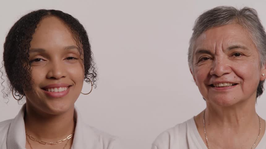 Two women of different generations a younger woman with curly hair and an older woman with short gray hair smile warmly at the camera they stand close together against a light background their expressions conveying a genuine and joyful connection