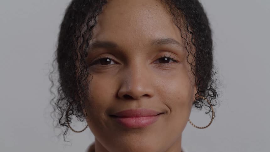 A closeup shot of a woman with dark curly hair and large hoop earrings shows her transition from a neutral expression to a gentle smile the woman is centered against a light gray background looking directly at the camera