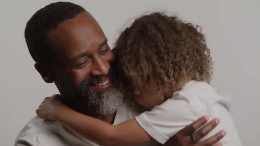 A father with a saltandpepper beard is shown in a close embrace with a young child who has curly hair they are both wearing white tops and the father smiles warmly down at the childs head